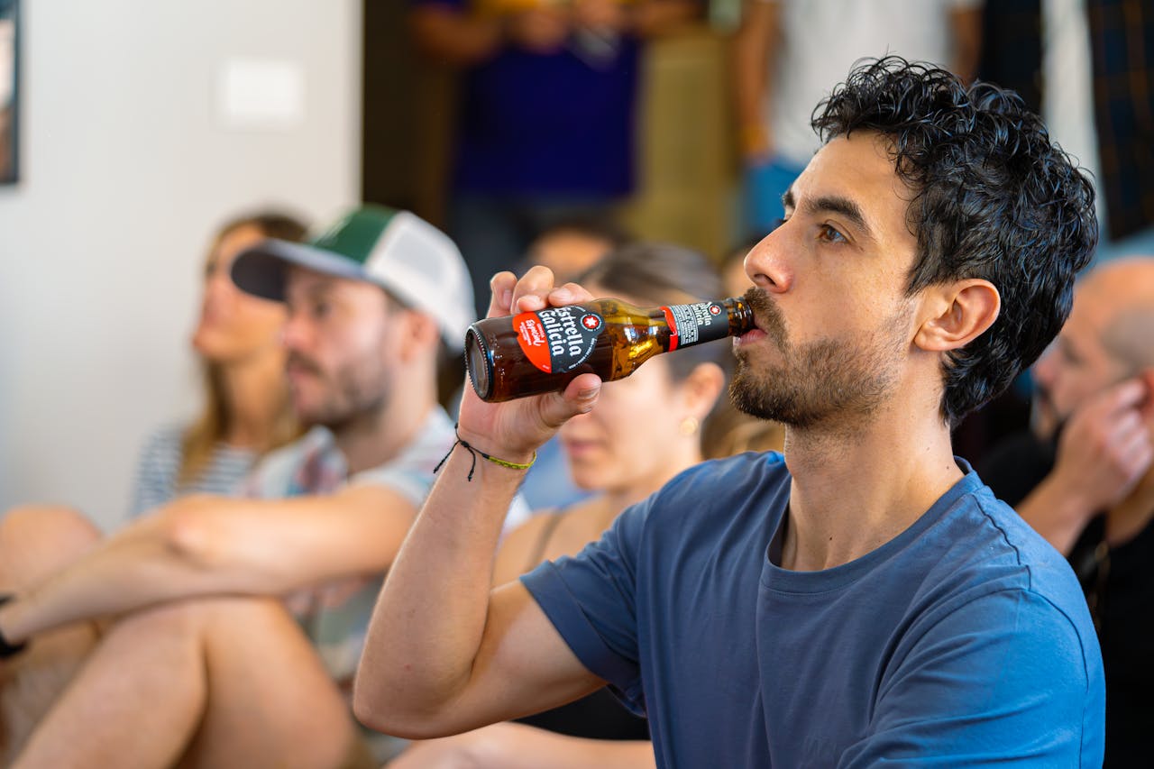 Close-up of a young man drinking Estrella Galicia beer at a casual indoor gathering.