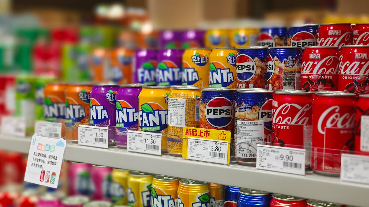 Colorful soda cans including Fanta, Pepsi, and Coca-Cola arranged on supermarket shelves, price tags visible.